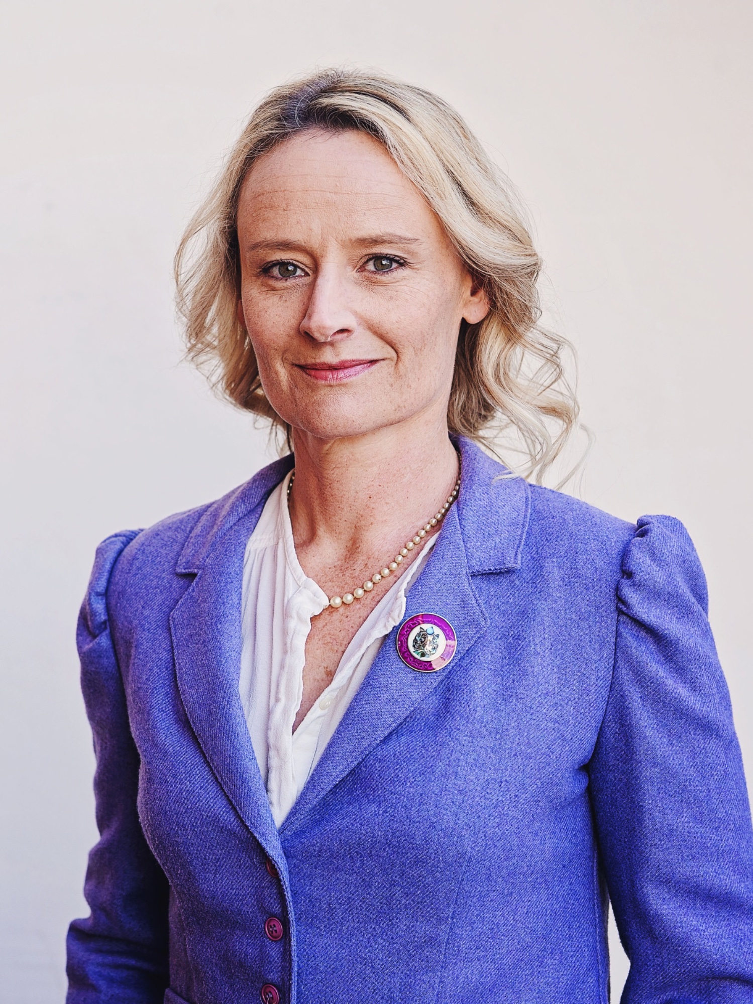 Head-and-shoulders portrait of Sarah McVie, wavy blonde hair, wearing a purple blazer and white blouse.