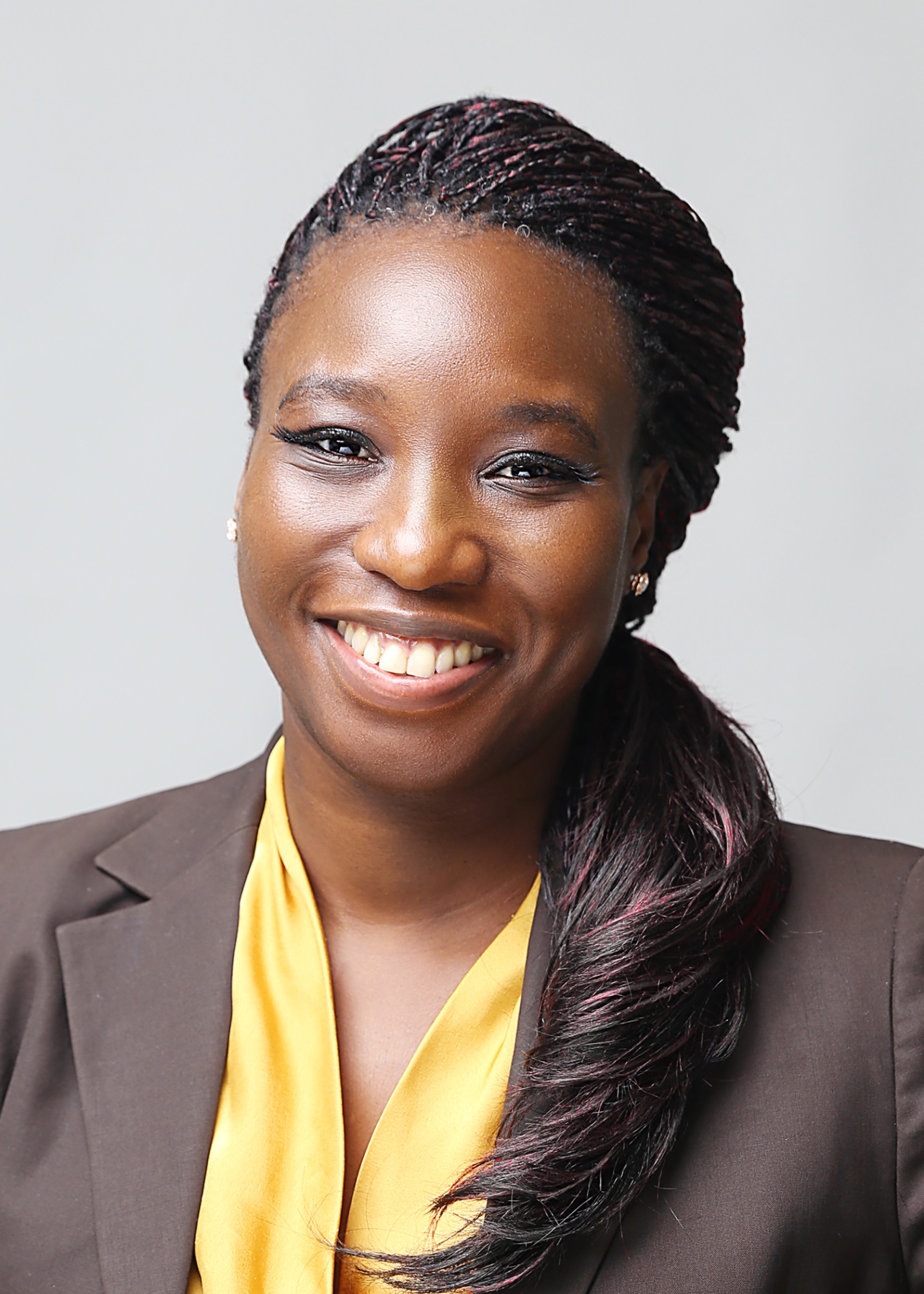 Head-and-shoulders portrait of Angela Ogang, long braided hair, wearing a brown blazer over a yellow top.
