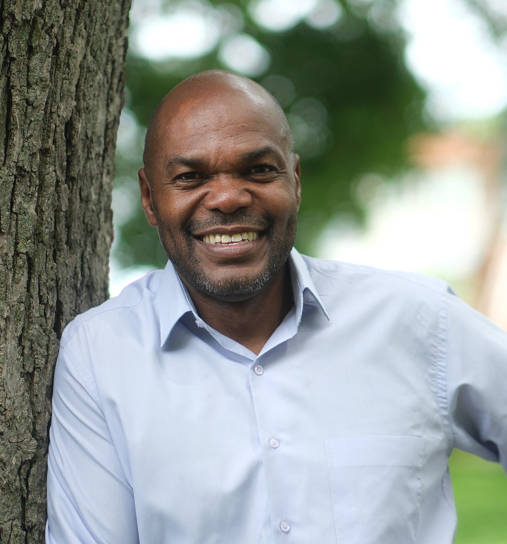 Head-and-shoulders portrait of Chris Moise, bald, with light stubble, wearing a light blue button-up shirt.