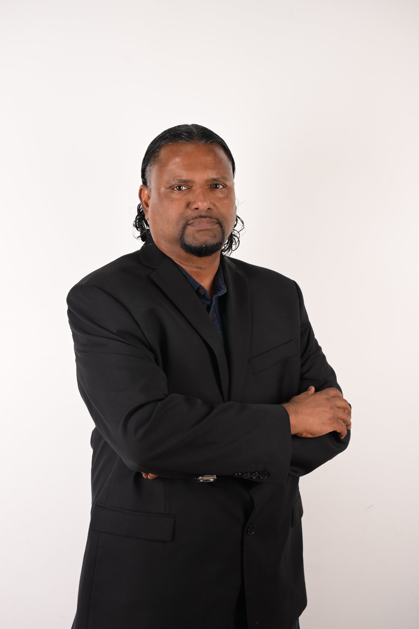 Head-and-shoulders portrait of Anton Perera, dark hair tied back and a goatee, wearing a black suit.