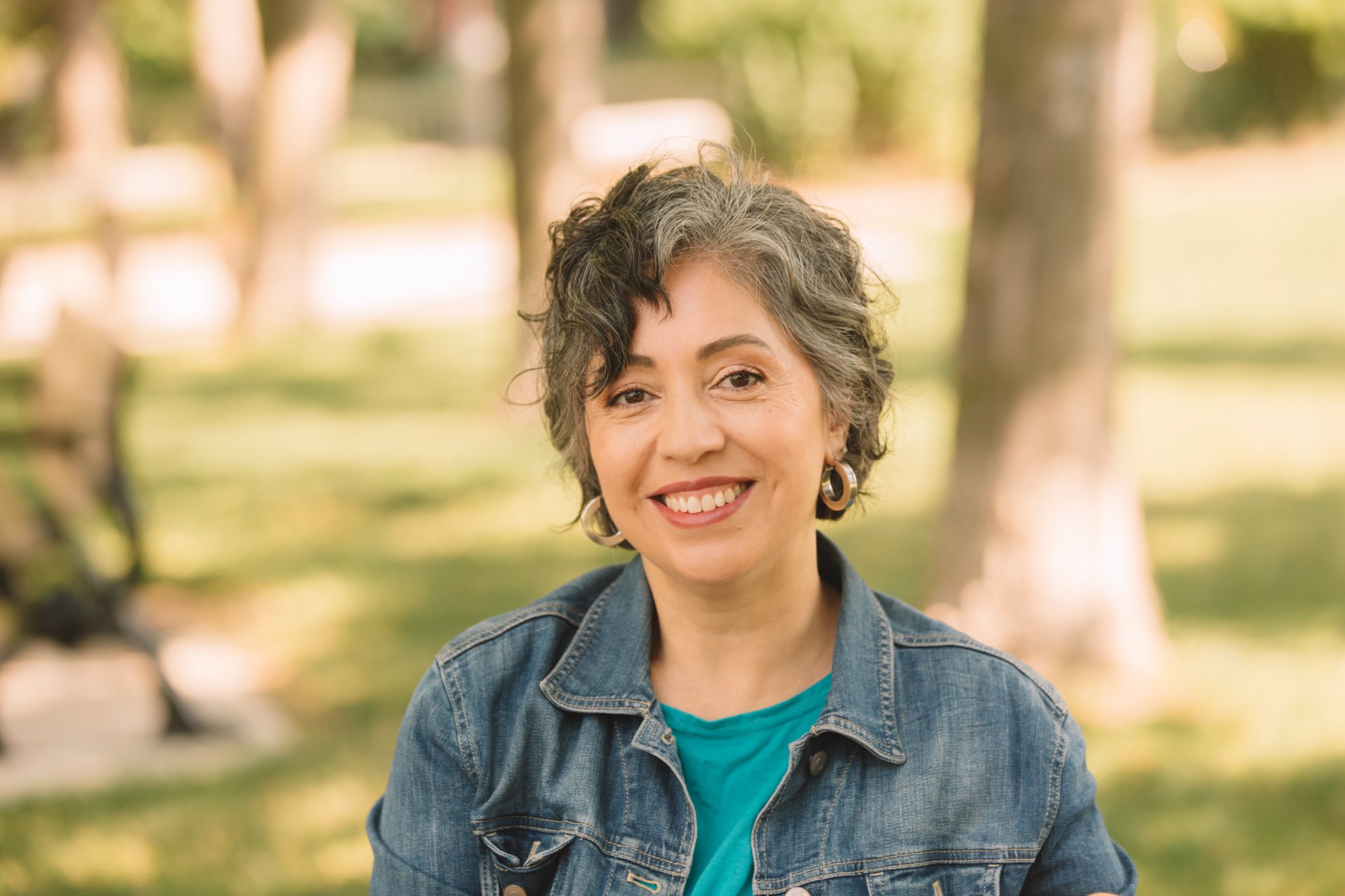 Head-and-shoulders portrait of Alejandra Bravo, short curly grey hair, wearing a teal top and a denim jacket.