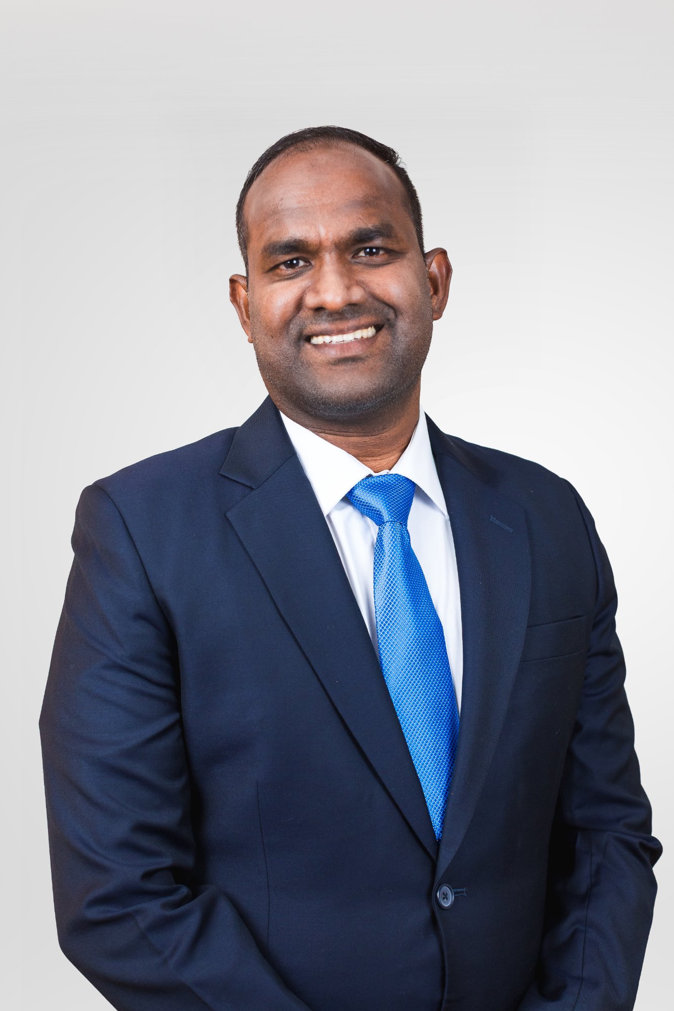 Head-and-shoulders portrait of Hassan Mubarak Noor Mohamed, short dark hair and light stubble, wearing a navy suit, white shirt, and blue tie.
