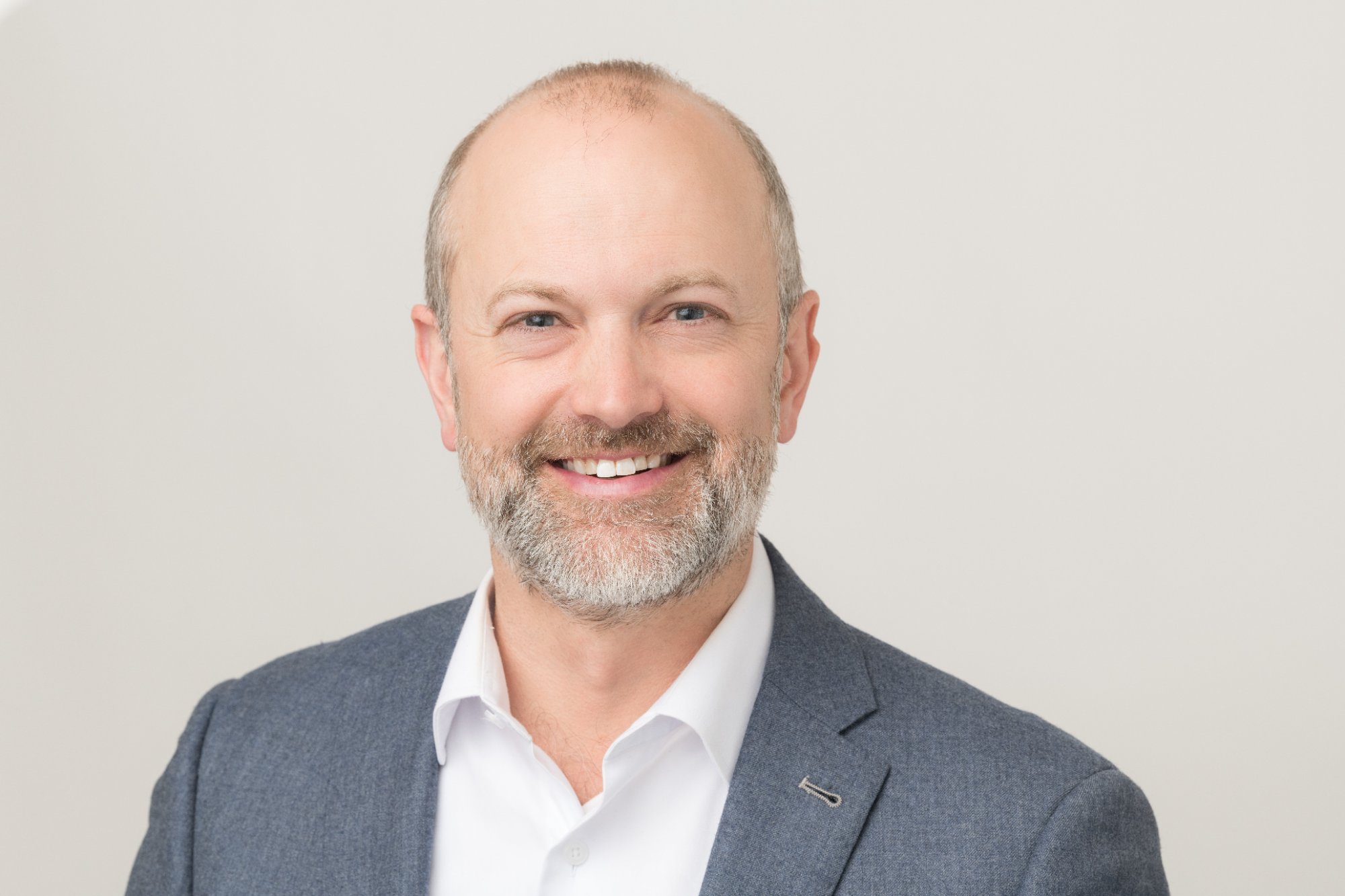 Head-and-shoulders portrait of Mike Layton, short grey hair and a grey beard, wearing a grey blazer and white shirt.
