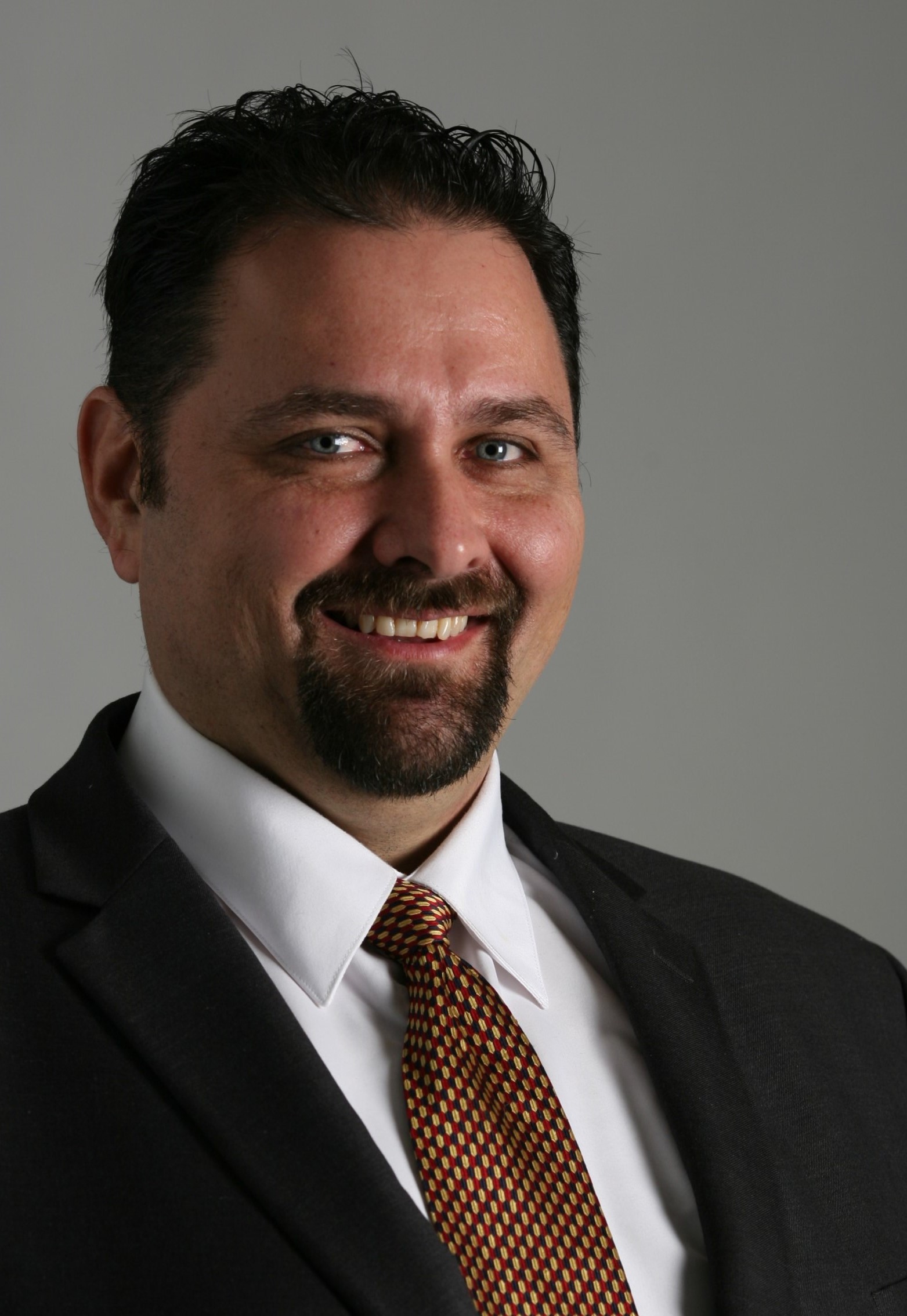 Head-and-shoulders portrait of Enzo Torrone, short dark hair and a beard, wearing a dark suit and patterned tie.