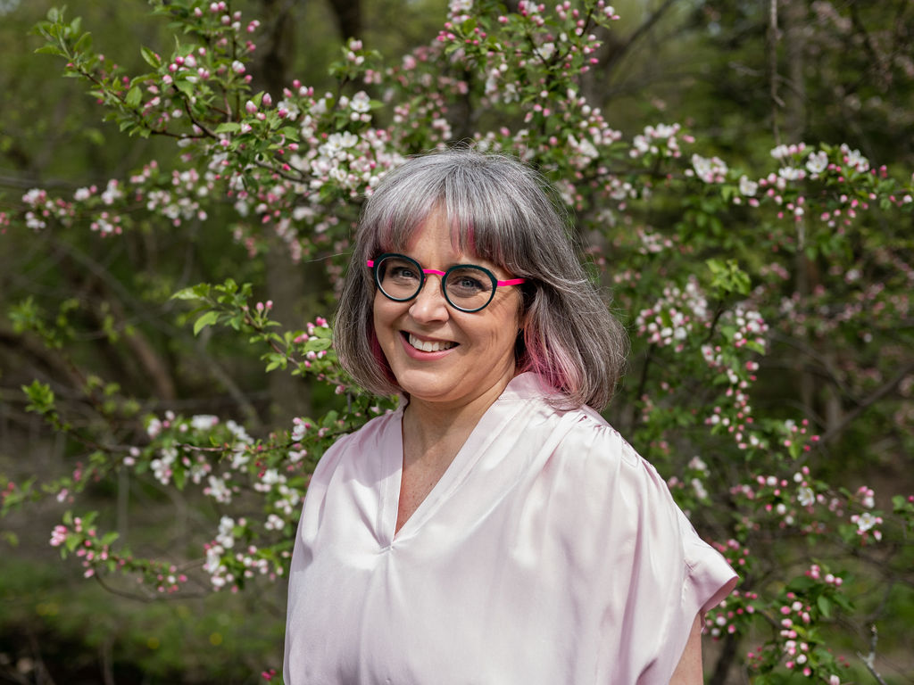 Head-and-shoulders portrait of Jennie Worden, grey hair with pink tips, wearing a light pink top and glasses.