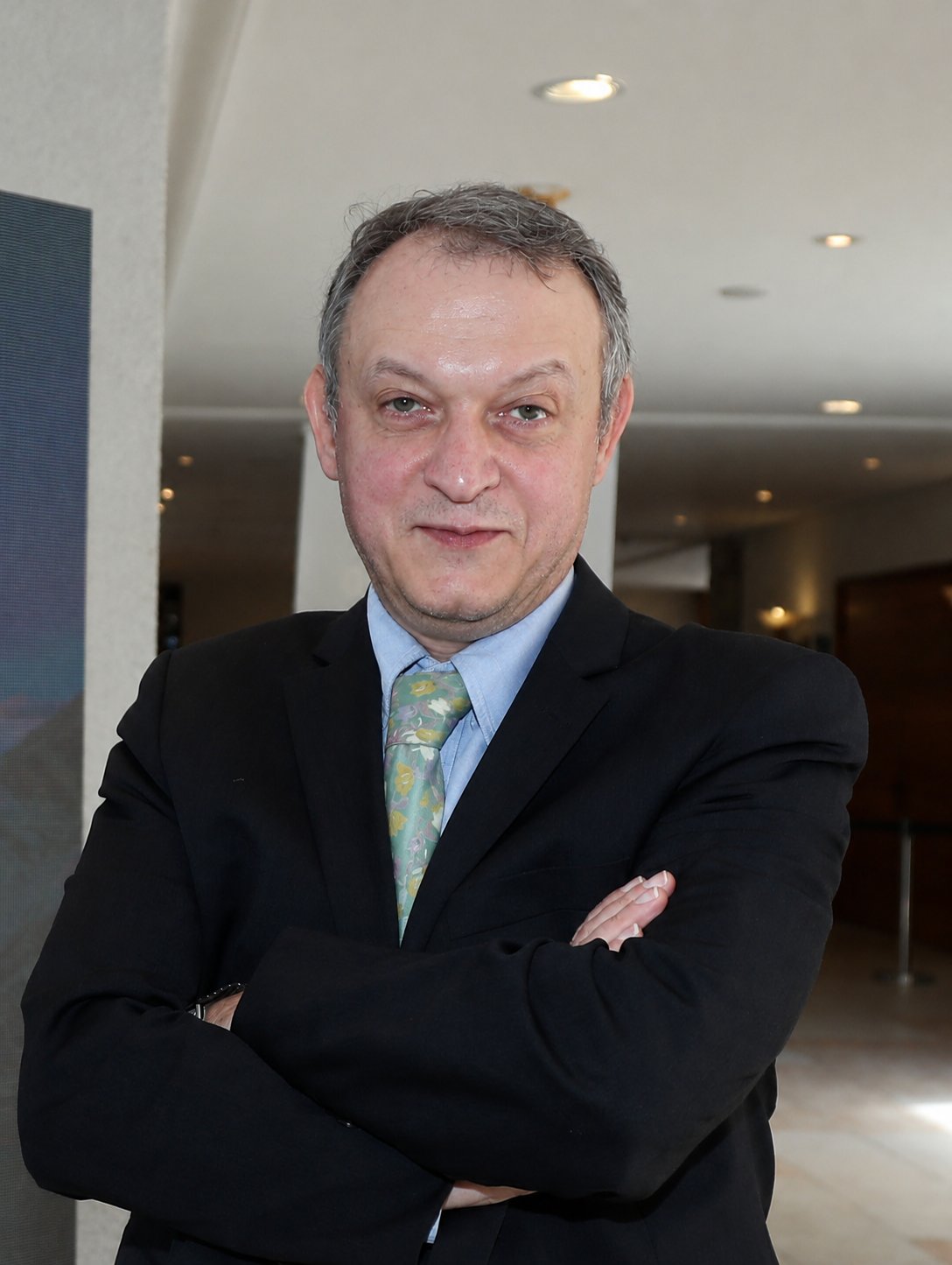 Head-and-shoulders portrait of Bashar Kassir, short grey hair, wearing a black suit and patterned tie.