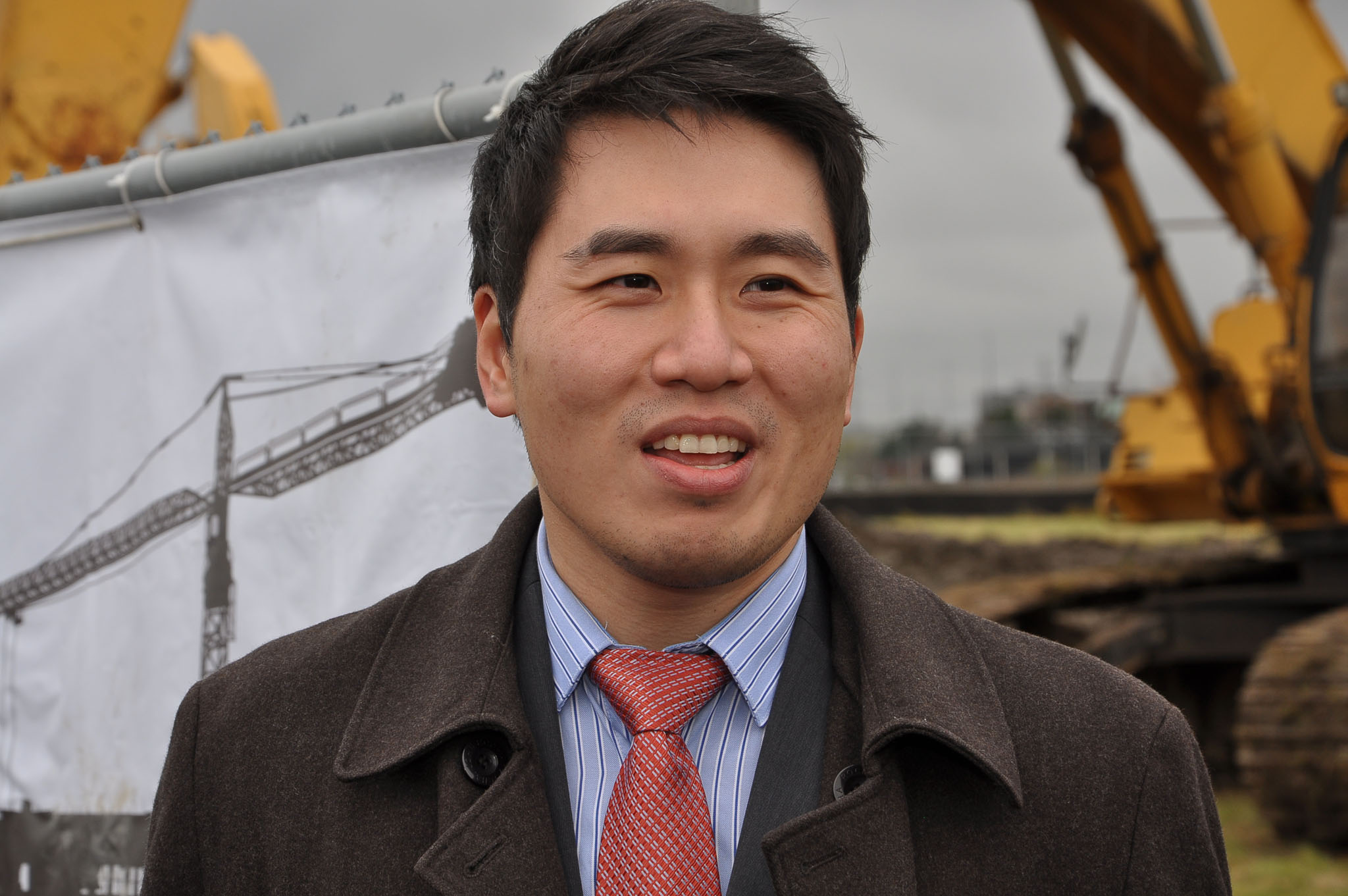 Head-and-shoulders portrait of Shaun Chen, short dark hair and light stubble, wearing a brown coat, striped shirt, and red tie.