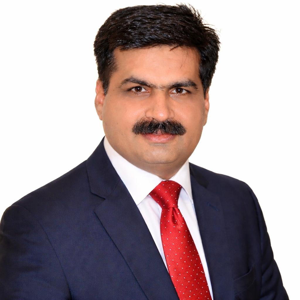 Head-and-shoulders portrait of Maqsood Ahmad, short dark hair and a moustache, wearing a navy suit, white shirt and red tie.